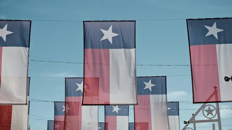 Multiple Texas state flags hanging in rows against a clear blue sky at an outdoor venue.